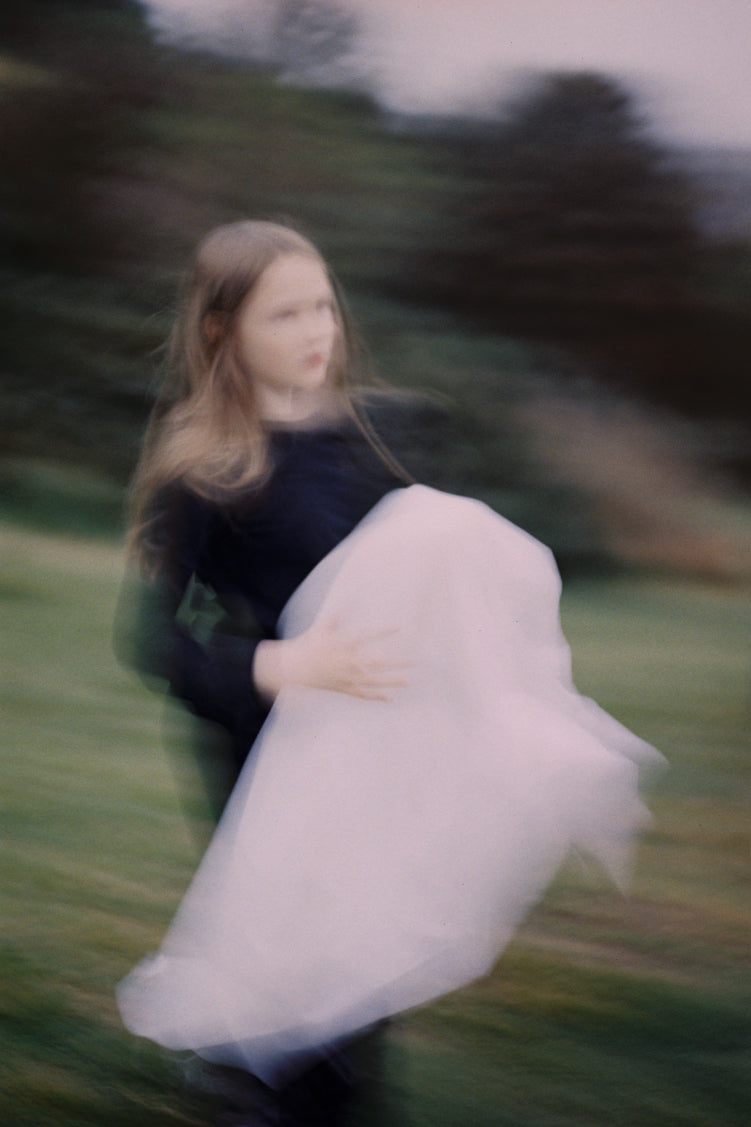 ALT= A photograph taken at the Wings workshop. Photo shows a young girl holding a bundle of white fabric. The image is blurred, and in the background are bushes, grass, and trees.