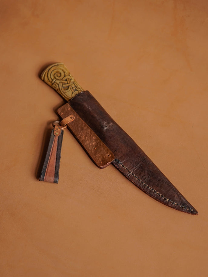ALT=The blade is sheathed in a hand-stitched and tooled scabbard of light cowhide, reinforced with hammered copper placquets and copper suspension rings.