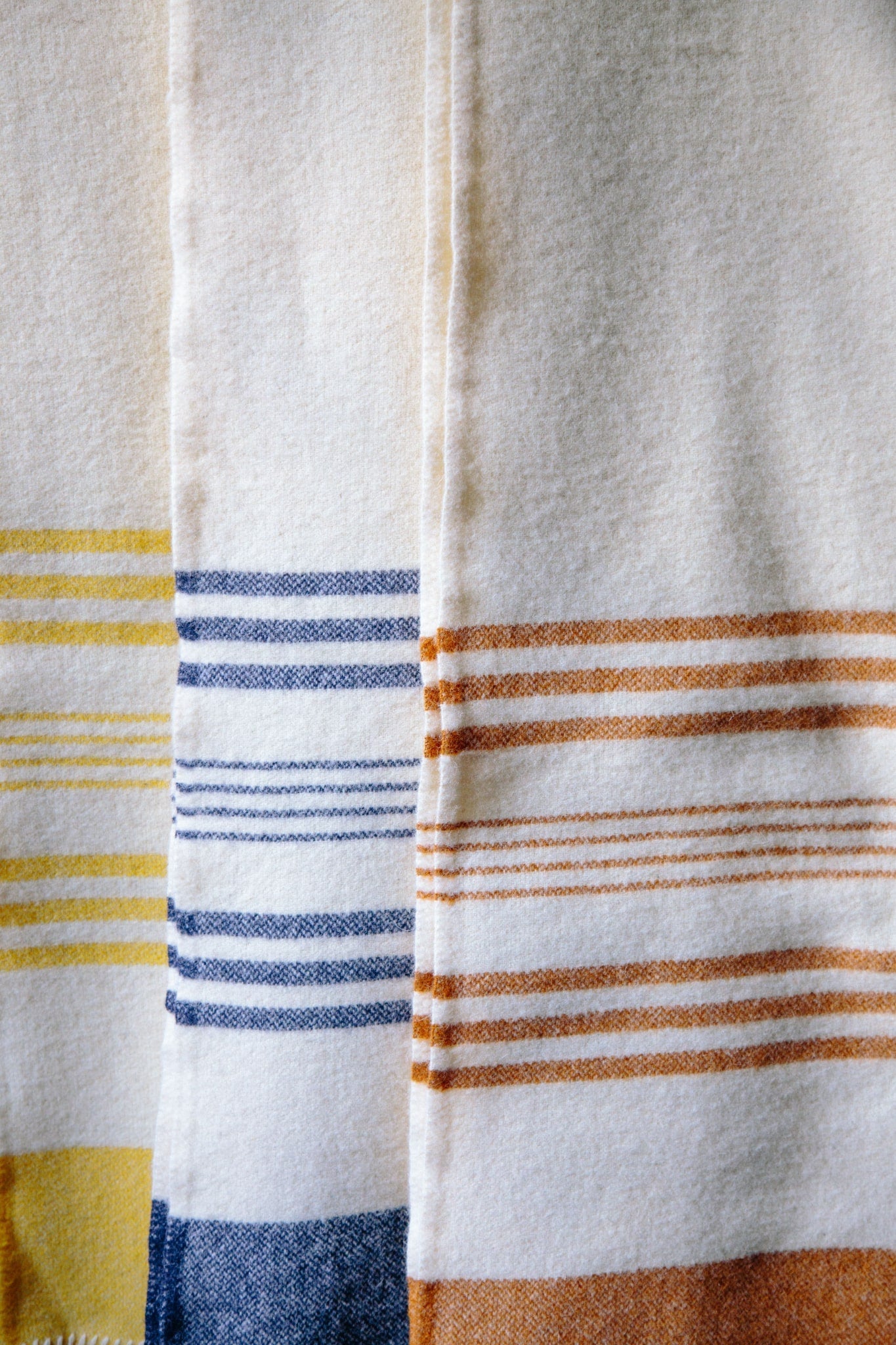 ALT=Three striped lambswool blankets in yellow, ochre and navy blue, hung vertically infront of eachother. The thick, weight of the blankets can be seen.
