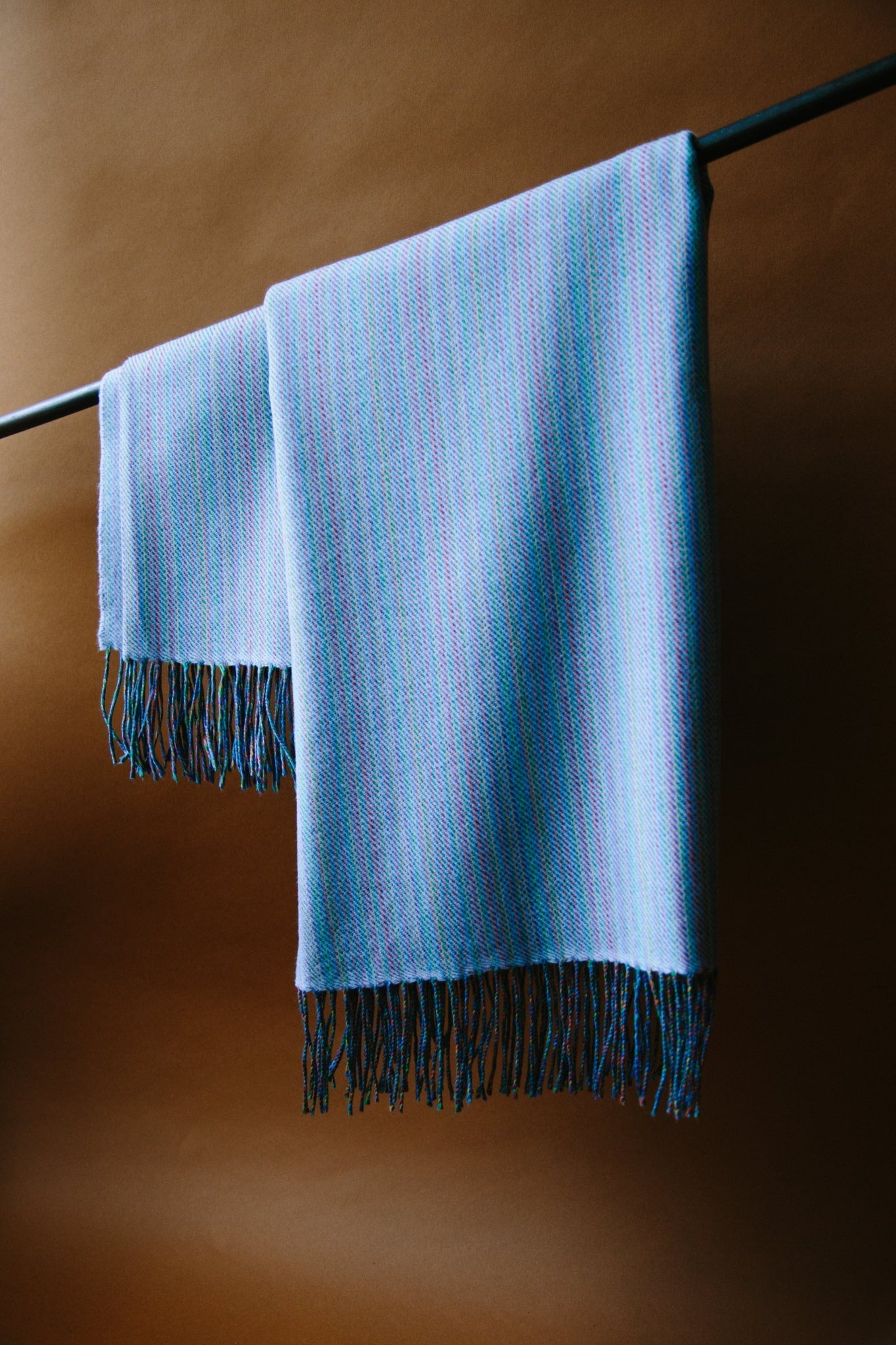 ALT=A sky blue merino wool throw hung from a black curtain pole. The throw has tassels at each end and is reflecting the natural light from a window on the left of the shot.