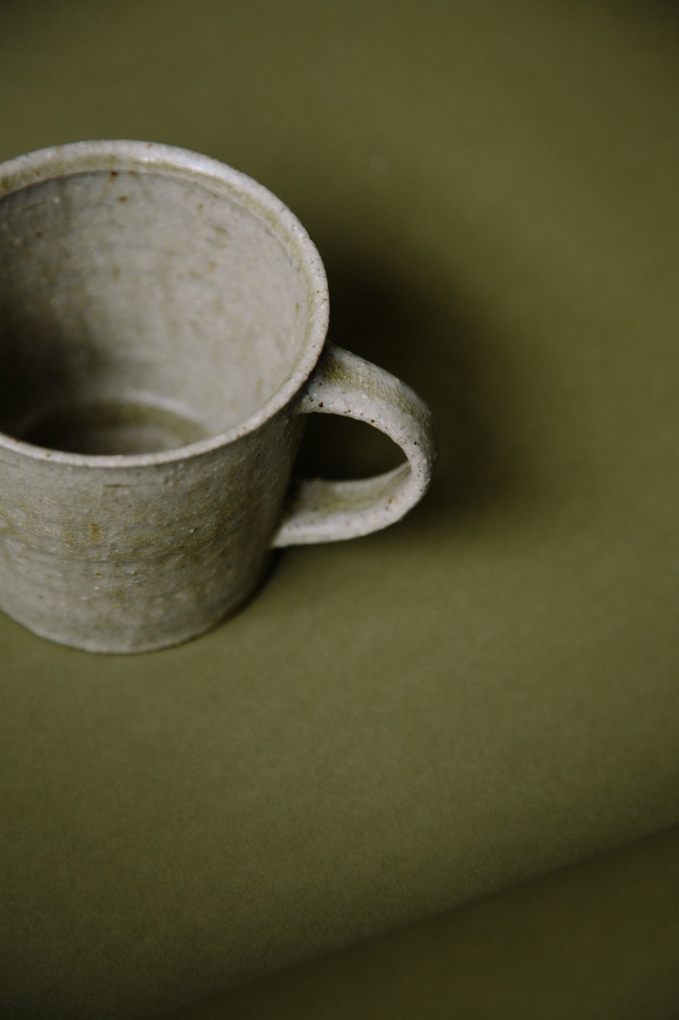ALT=An ash-glazed hand-thrown ceramic mug in a grey-green neutral shade with a curved handle. Photographed on a olive green background from above so the inside of the mug can be seen.