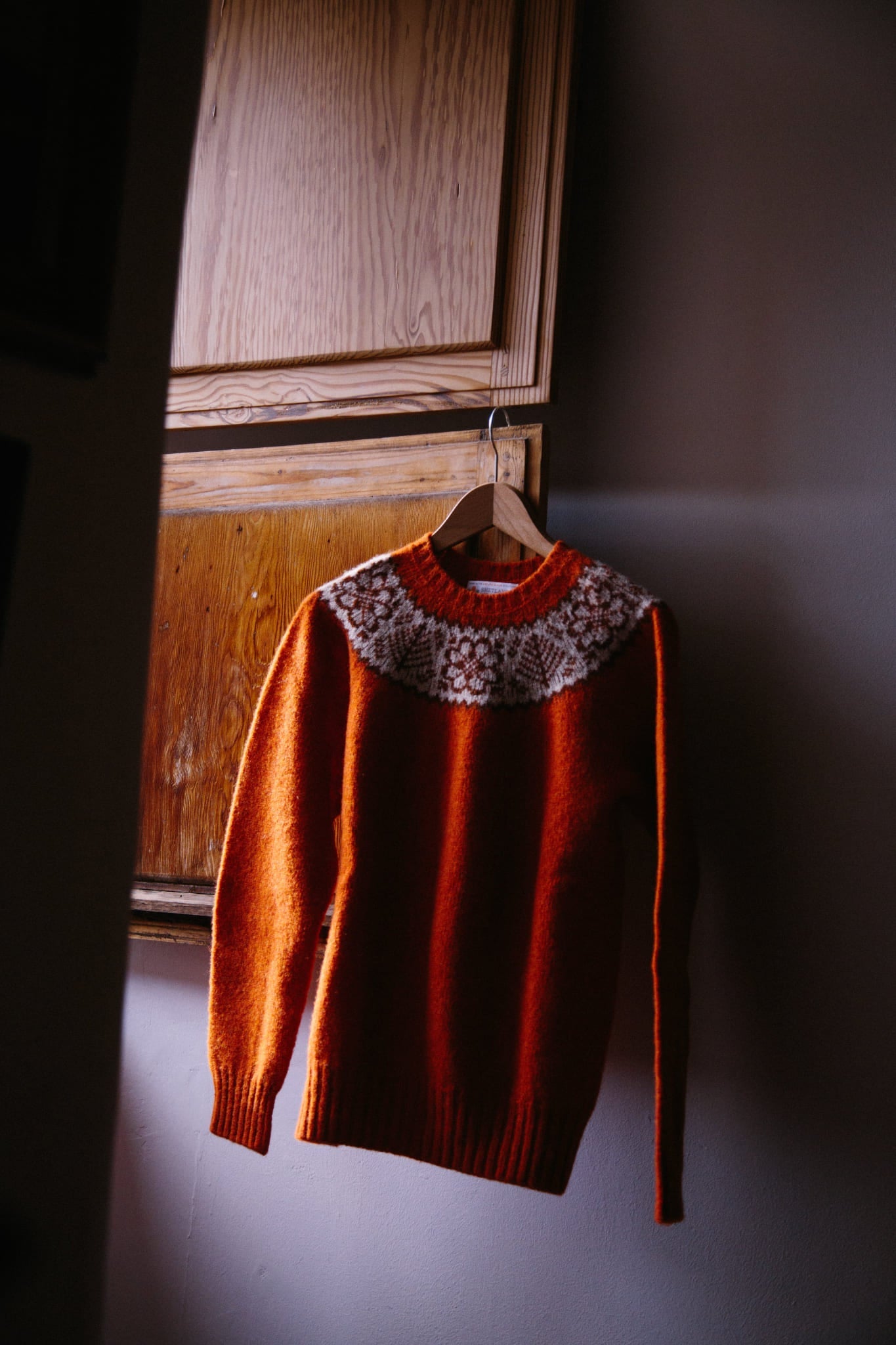 ALT=A Shetland woolen pullover jumper in Fair Isle Yoke, iron rust red. Hung on a wooden hanger from a window shutter.