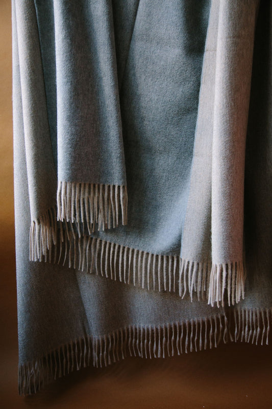 ALT=Large cashmere throw. Two sided reversible colour; grey and pale beige. Draped against a brown background with natural light.