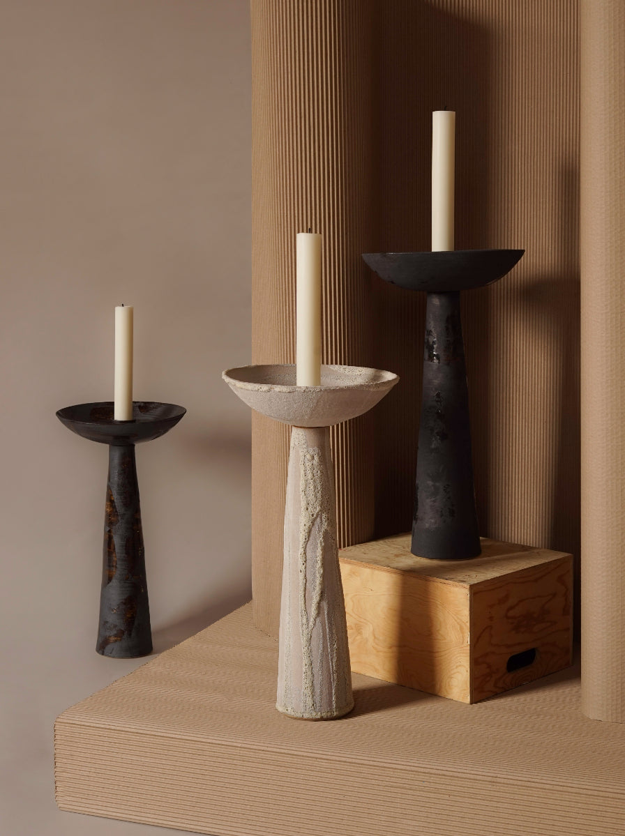 ALT= A row of three ceramic candlesticks holding white church candles with burnt wicks. On the left is a black and copper candlestick, the middle a soft grey, and the right, black. They have been photographed amongst packaging materials. 