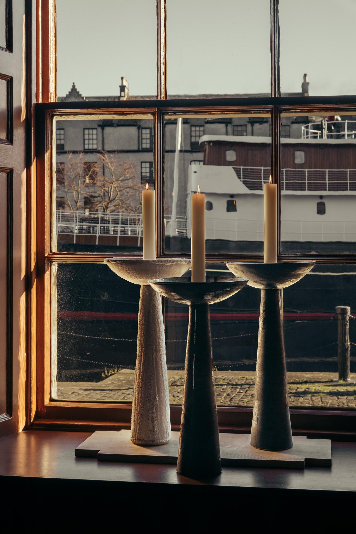 ALT= Three Samhain Candlesticks by ceramicist Cara Guthrie. Shown in each of the three colours; Slate, Iron and Lichen. Each candlestick stands tall and holds a white candle set alight.