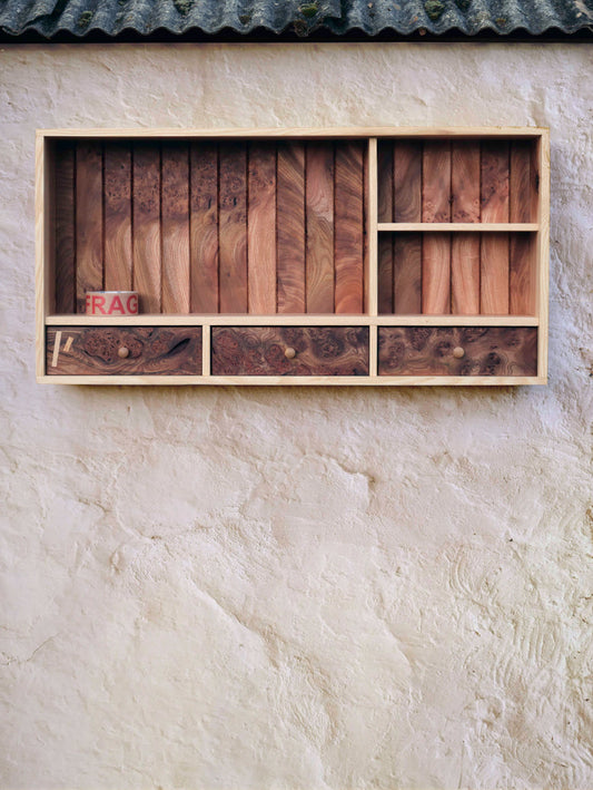 ALT= Tom Addy's Prìseil, is a wooden wall mounted storage shelf with three drawers. Photographed here outdoors, on a textured beige wall, with the weathered guttering visible at the very top of the image.