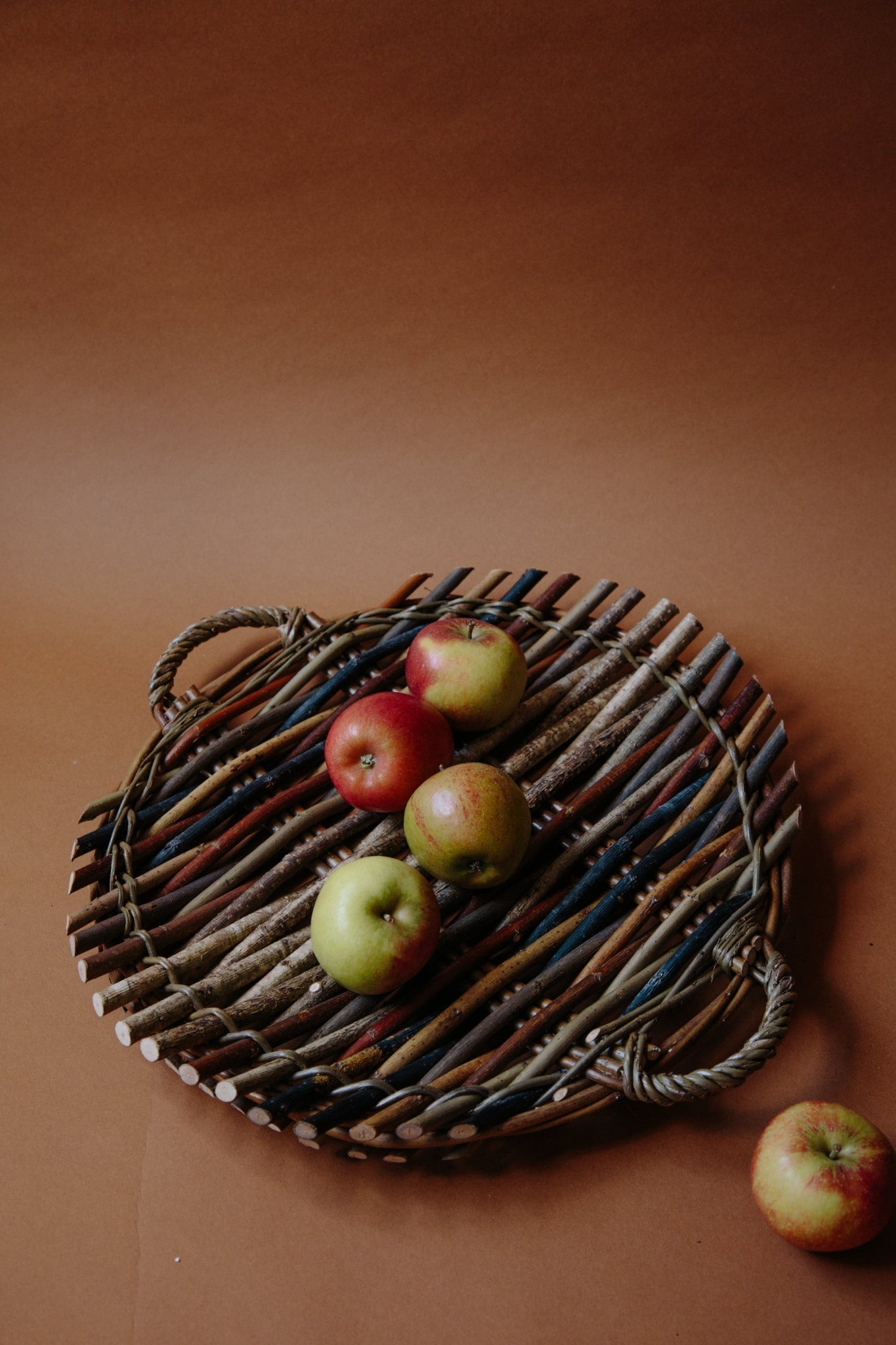 ALT=Circular willow platter woven from woods and willow with two small handles on either side. Platter holds four apples, with a fifth rolling out of frame. Photograph shot on a deep brown background.