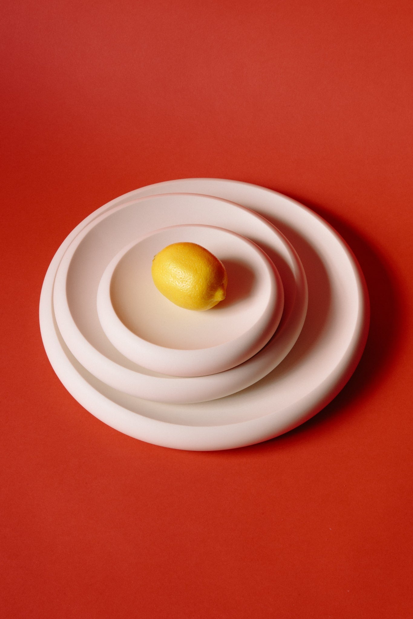 ALT=Three white jesmonite plates on a red background. Stacked in size order, small, medium, and large. A lemon is on the smallest plate. 