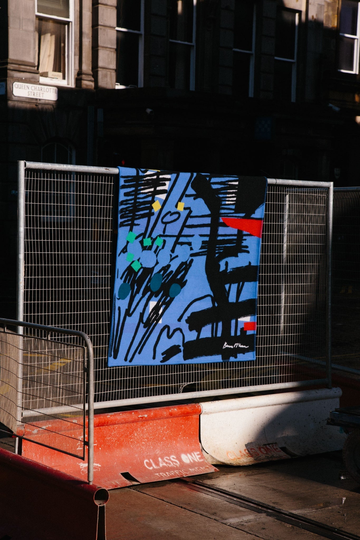 ALT=Large woven lambswool and cashmere blanket in blues, black, reds, greens and yellow with graphic hand-drawn lines that have been translated using a jacquard loom. Blanket laid over a traffic works fence in Leith, Edinburgh. 