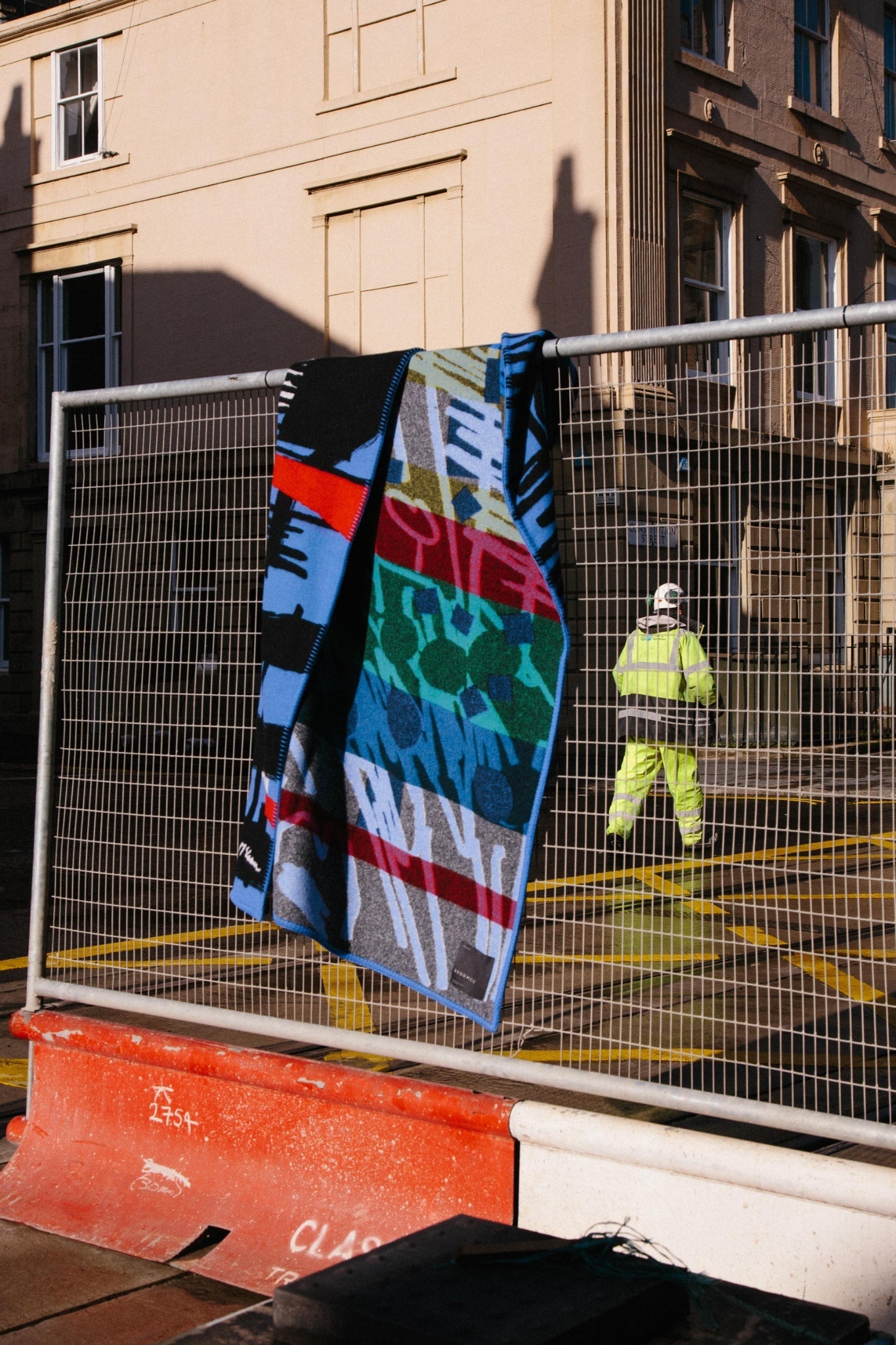 ALT=Large woven lambswool and cashmere blanket in blues, black, reds, greens and yellow with graphic hand-drawn lines that have been translated using a jacquard loom. Blanket laid over a traffic works fence in Leith, Edinburgh. with a worked in high-viz in the background.