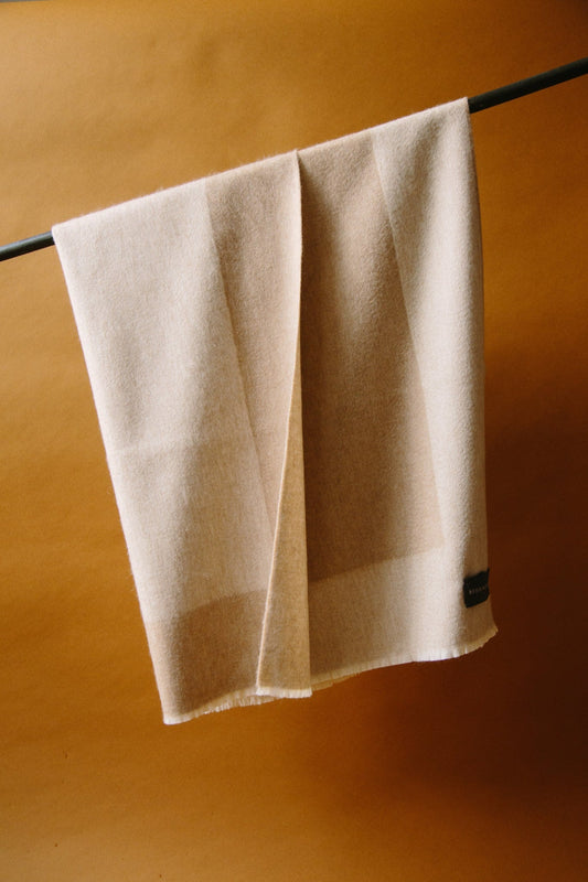 ALT=Two colour cashmere baby blanket in white and beige. Draped over a black curtain pole against a brown paper background with natural light.