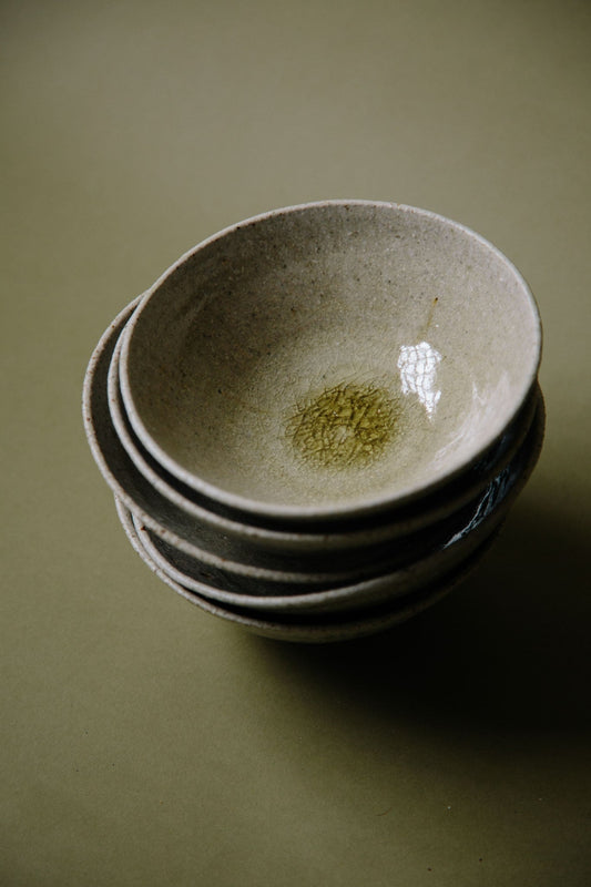 ALT=Glossy glazed bowls in neutral green and grey shades with subtle crackling glaze in the centre of the bowl. Photographed in a stack of five on an olive green background.