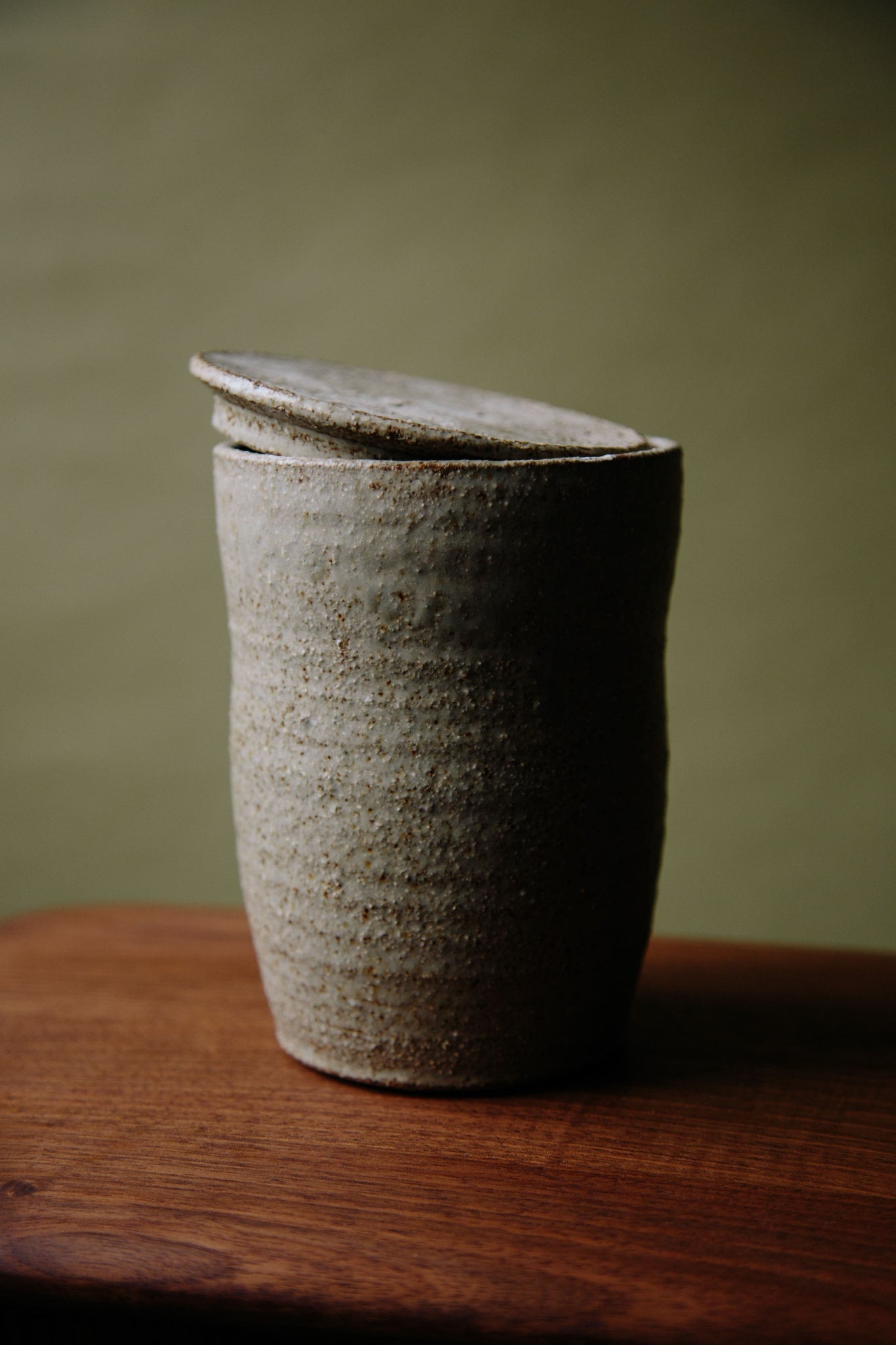 ALT=A textured, hand-thrown ceramic jar with a lid photographed on a wooden stool against an olive green background. The lid is at an angle so the shape can be seen.