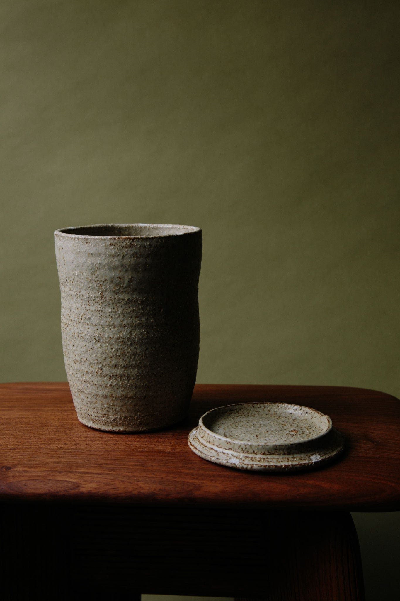 ALT=A textured ceramic jar with a lid photographed on a wooden stool against an olive green background. The lid has been taken off and placed next to the jar.