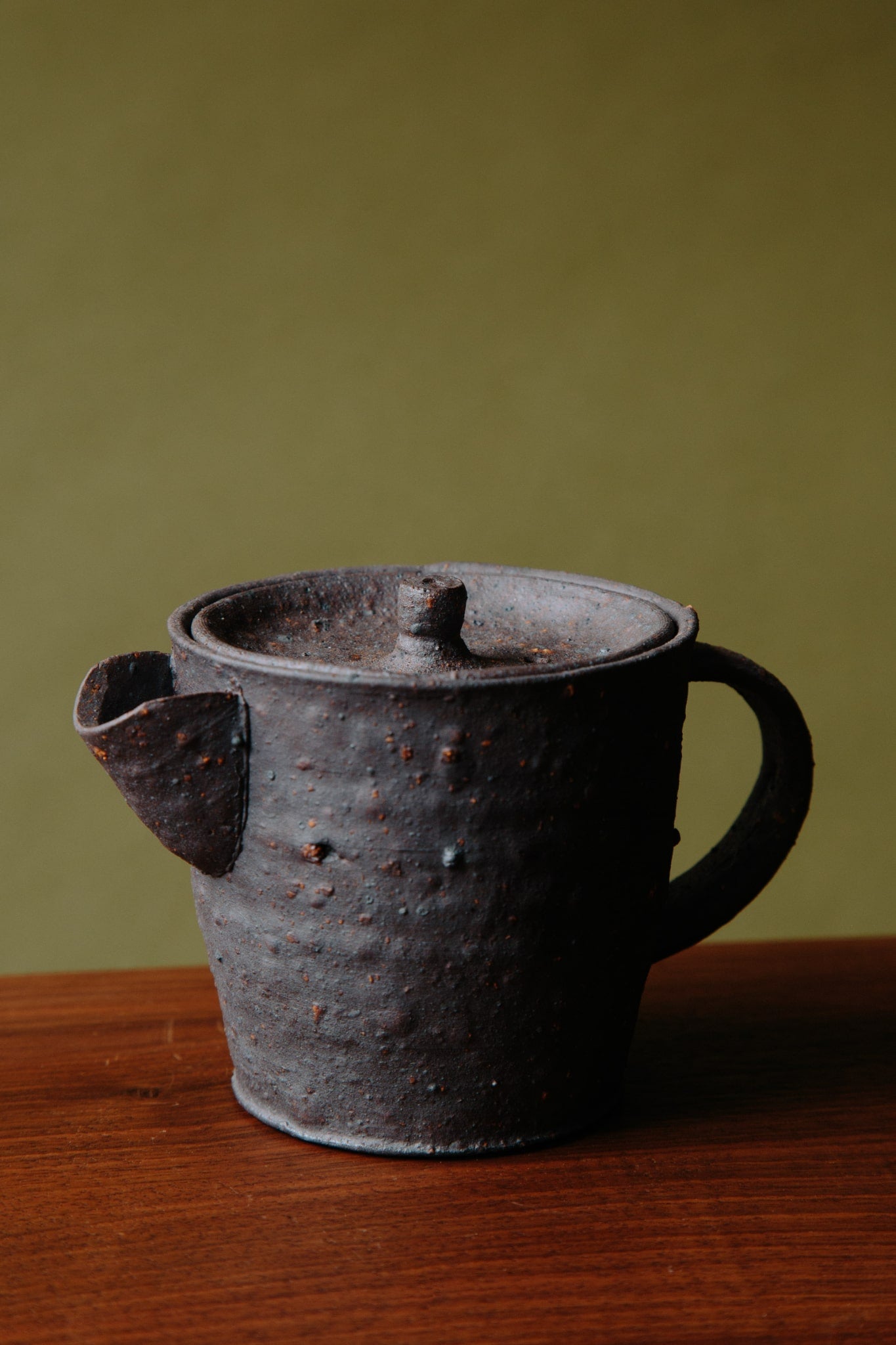 ALT=A small black ceramic teapot with a characterful spout, lid and handle. Shot on a wooden stool against a green background.