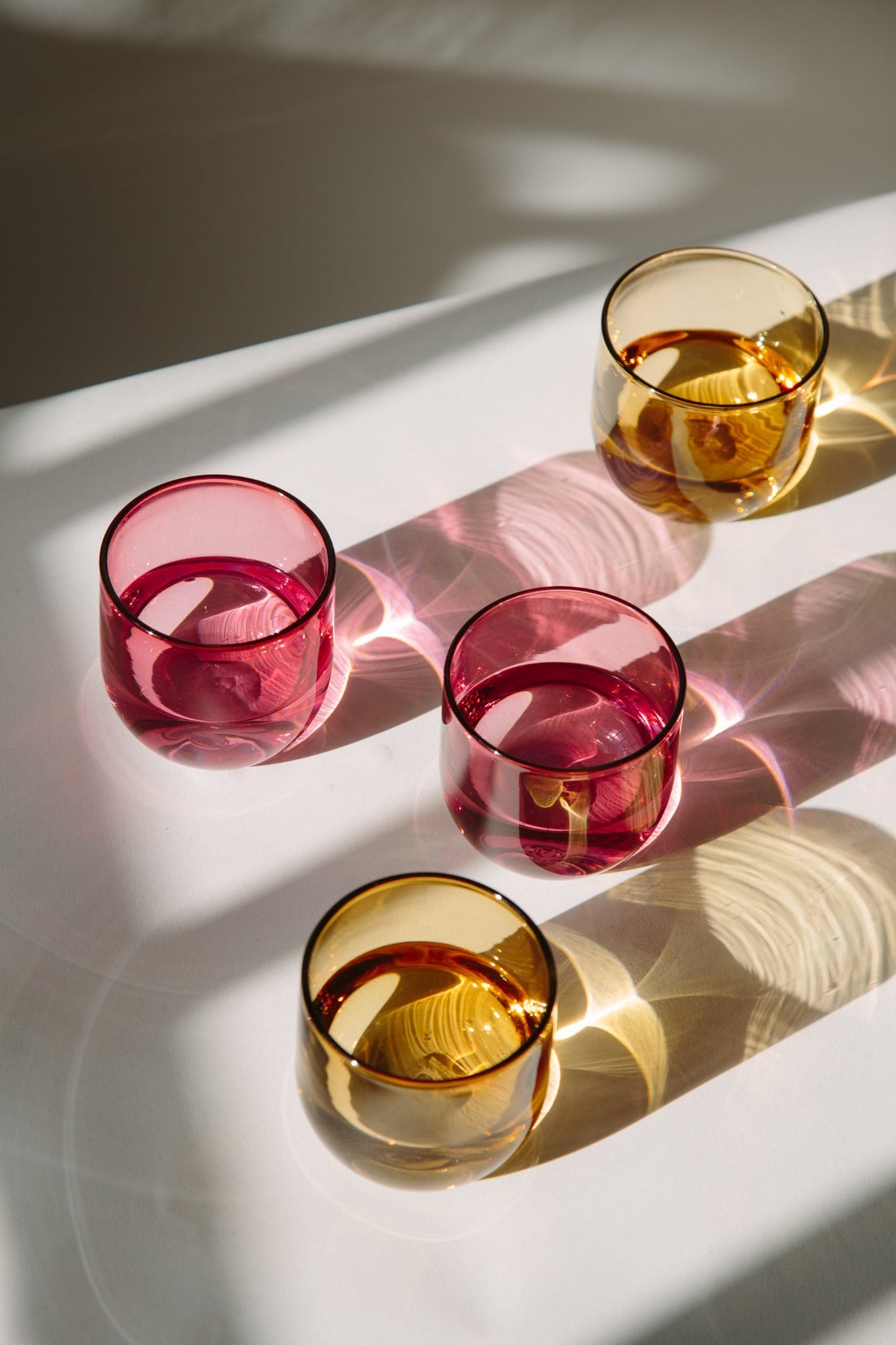 ALT=Four solid coloured class drinking cups filled with water, two pink and two amber. Colourful reflections cover the white paper suface underneath.