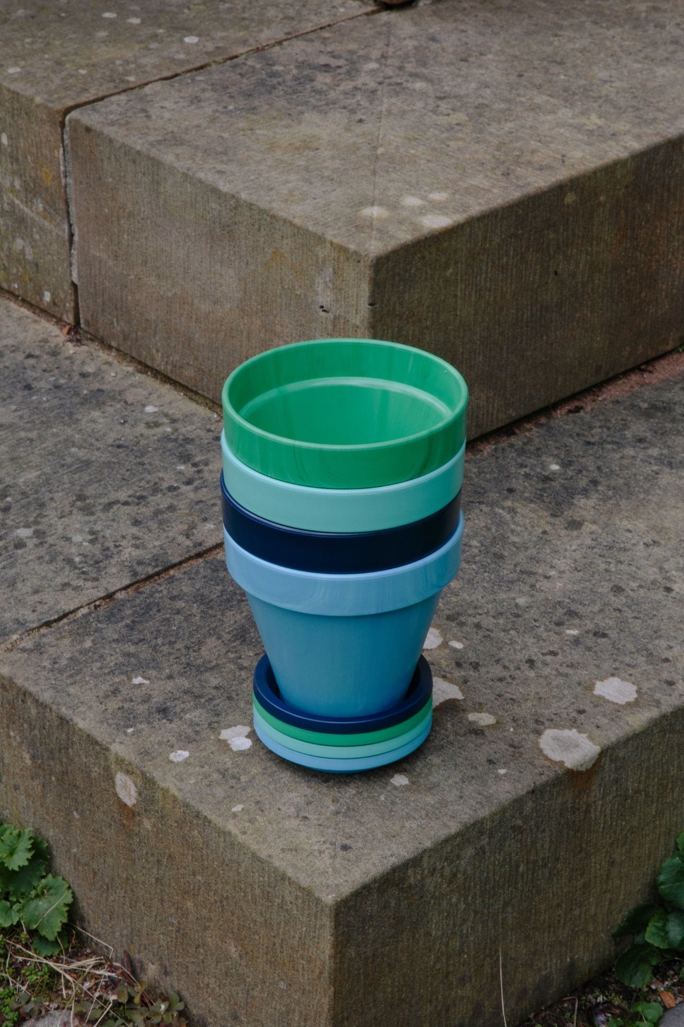 ALT=A stack of green and blue recycled plastic plant pots with saucers. On the edge of a set of stone steps.