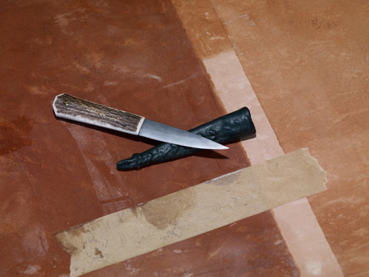 ALT=An Antler Sgian Dubh knife balanced on top of its handcrafted leather sheath. Shot on a plaster background.